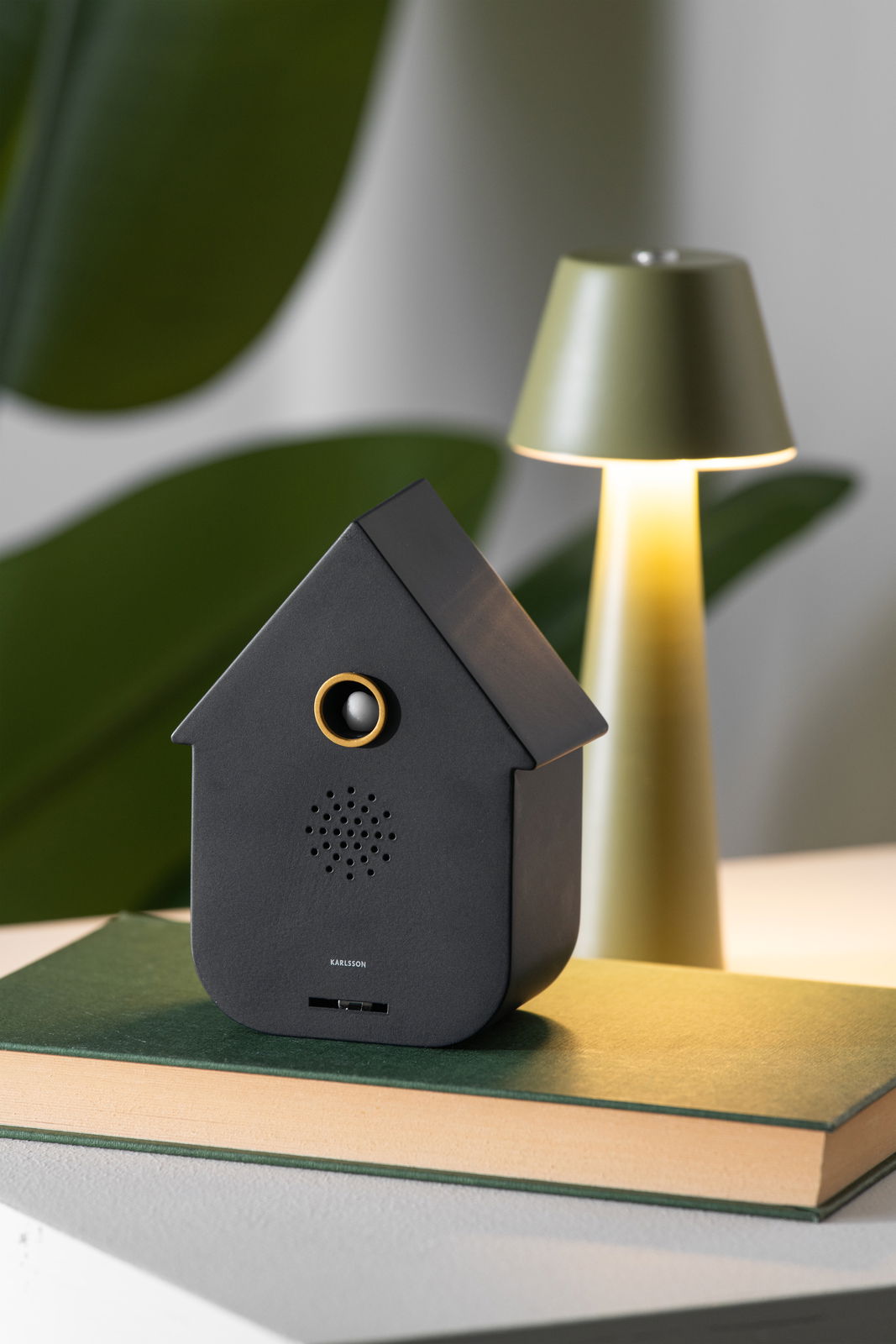 KARLSSON Soundbox CUCKOO Schwarze, moderne Soundbox in Form eines Hauses, frontal auf einem Buch platziert, mit einer Lampe im Hintergrund.