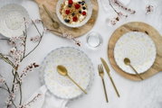 Draufsicht auf dekoriertes Geschirr-Set mit pastellfarbenen Blumenmustern auf weißem Porzellan, goldfarbenem Besteck und Schalen mit Joghurt, Granola und Beeren auf Holzuntersetzern, umgeben von Blütenzweigen.