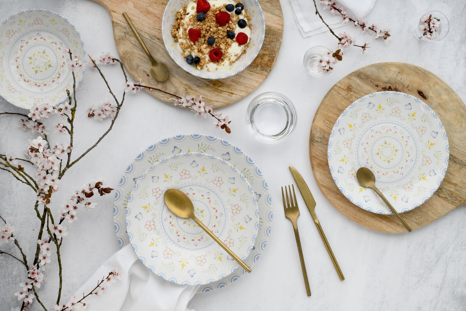 Draufsicht auf dekoriertes Geschirr-Set mit pastellfarbenen Blumenmustern auf weißem Porzellan, goldfarbenem Besteck und Schalen mit Joghurt, Granola und Beeren auf Holzuntersetzern, umgeben von Blütenzweigen.