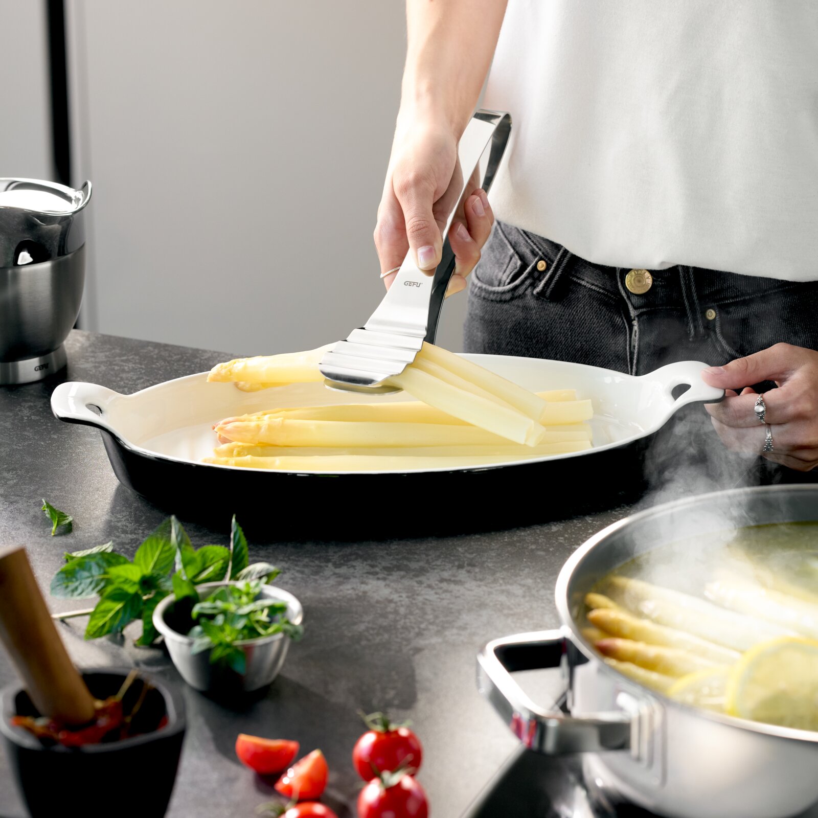 Schwarze ovale Auflaufform mit weißen Griffen, in der eine Person mit Edelstahl-Küchenzange gekochten Spargel anrichtet; Perspektive: seitliche Nahaufnahme auf Arbeitsplattenhöhe.