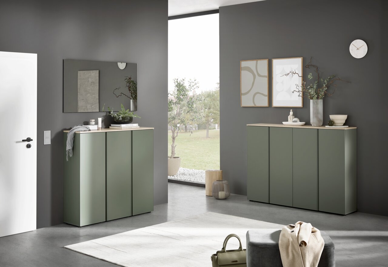 Voleo XXL-Schuhschrank MILTON Modernes Sideboard in mattem Salbeigrün mit heller Holzplatte in einem minimalistischen Wohnraum, frontale Perspektive mit leichtem Blick von links; dekoriert mit Pflanzen, Bildern und Spiegel an grauer Wand.