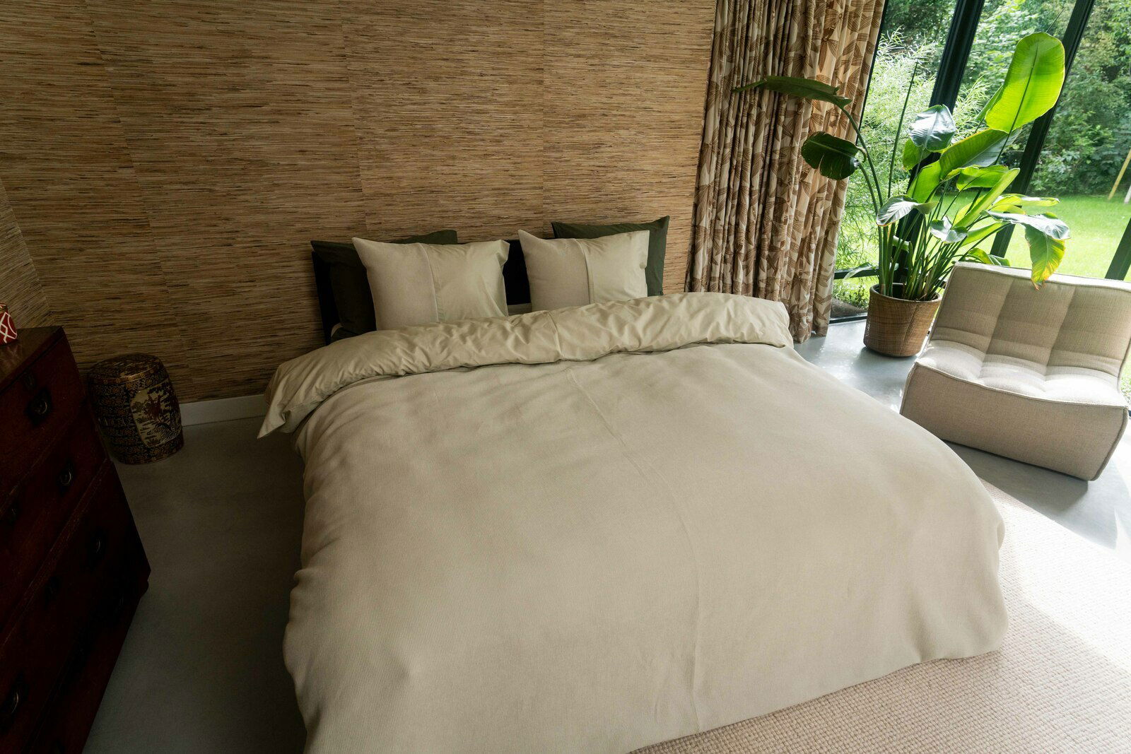 Gemütliches Schlafzimmer mit einem großen Bett, das mit beiger Bettwäsche bezogen ist. Im Hintergrund sind eine grüne Pflanze und ein großes Fenster zu sehen. Die Perspektive zeigt das Bett von vorne.