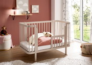 Weißes Babybett aus Holz mit Gitterseiten und kleiner seitlicher Einstiegstür, im Kinderzimmer neben bodentiefem Fenster; Perspektive: schräg von vorne (3/4-Ansicht).