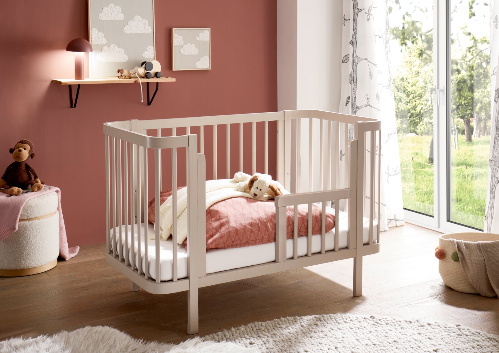 MyBaby Babybett SPICE/PEPPINI Weißes Babybett aus Holz mit Gitterseiten und kleiner seitlicher Einstiegstür, im Kinderzimmer neben bodentiefem Fenster; Perspektive: schräg von vorne (3/4-Ansicht).