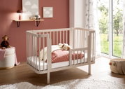 Weißes Babybett aus Holz mit Gitterseiten und kleiner seitlicher Einstiegstür, im Kinderzimmer neben bodentiefem Fenster; Perspektive: schräg von vorne (3/4-Ansicht).