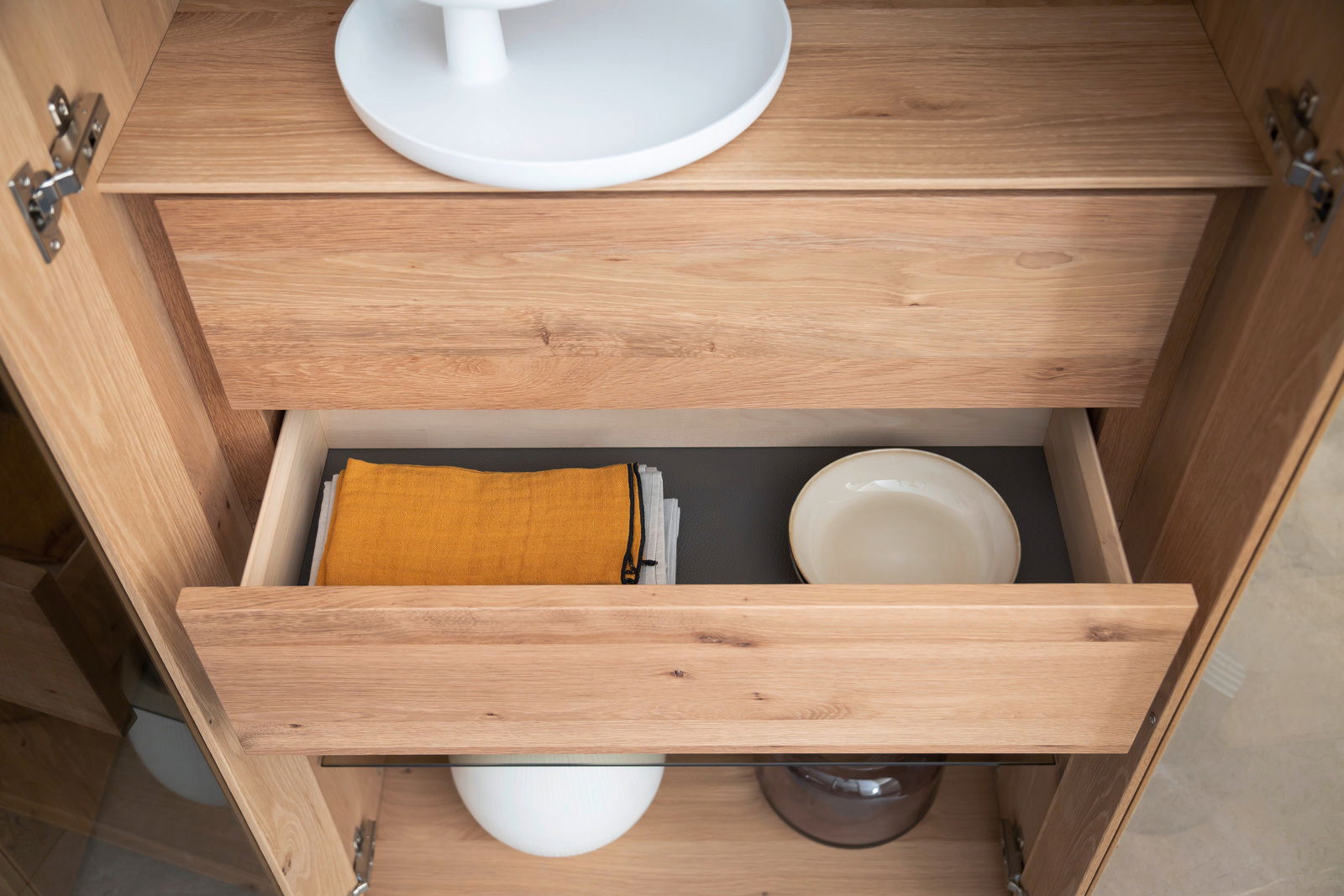 Geöffnete Schublade einer Vitrine aus Holz mit gelbem Tuch und weißem Teller, von oben betrachtet.