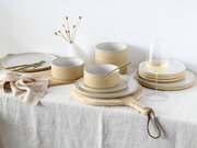Servies Futuro White Sand 12, bestaande uit elegante, zandkleurige borden en schalen, opgesteld op een tafel met een tafelkleed van natuurlijk linnen, gefotografeerd vanuit een zijdelingse hoek.