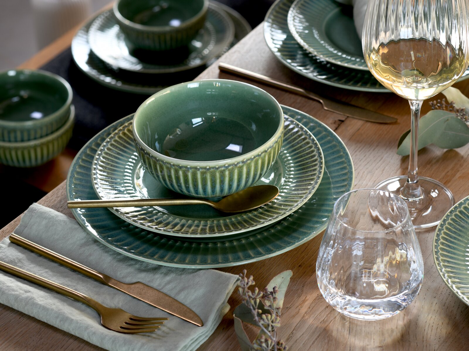 Schräg von oben fotografiertes Tischgedeck mit grün glasiertem Keramikgeschirr (Schale auf gerippten Tellern), goldenem Besteck und Weinglas auf Holztisch.