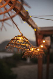 Nahaufnahme einer Outdoor-Solarlichterkette mit dekorativen Lampenschirmen aus Rattan, aufgehängt an einem Holzrahmen in der Abenddämmerung.