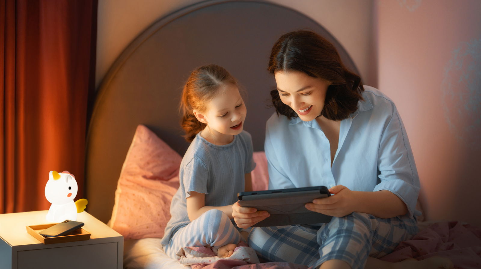 FHL easy Akku-Nachtleuchte UNICORNO Eine Akku-Nachtleuchte in Form eines Einhorns steht auf einem Nachttisch neben einem Bett, auf dem ein Kind und eine Frau gemeinsam ein Tablet betrachten. Die Szene ist aus einer seitlichen Perspektive aufgenommen.