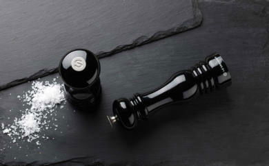 Schwarze glänzende Salzmühle, eine stehend und eine liegend, auf einer dunklen Schieferplatte mit grobem Salz daneben, aus der Vogelperspektive.