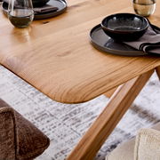 Nahaufnahme eines Esstisches aus Holz mit abgerundeten Ecken, gedeckt mit Geschirr und Gläsern, aus seitlicher Perspektive.