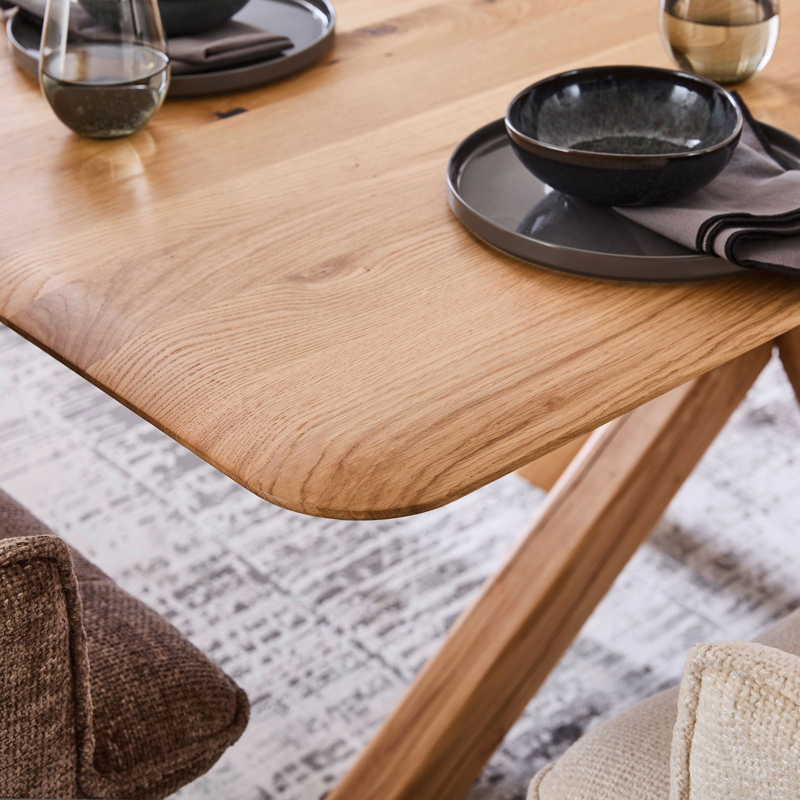 Nahaufnahme eines Esstisches aus Holz mit abgerundeten Ecken, gedeckt mit Geschirr und Gläsern, aus seitlicher Perspektive.