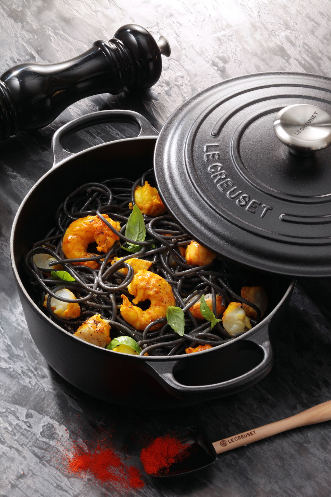 Schwarzer runder Le Creuset Bräter 26 cm mit Deckel, gefüllt mit Garnelen und schwarzer Pasta, aus der Vogelperspektive fotografiert.