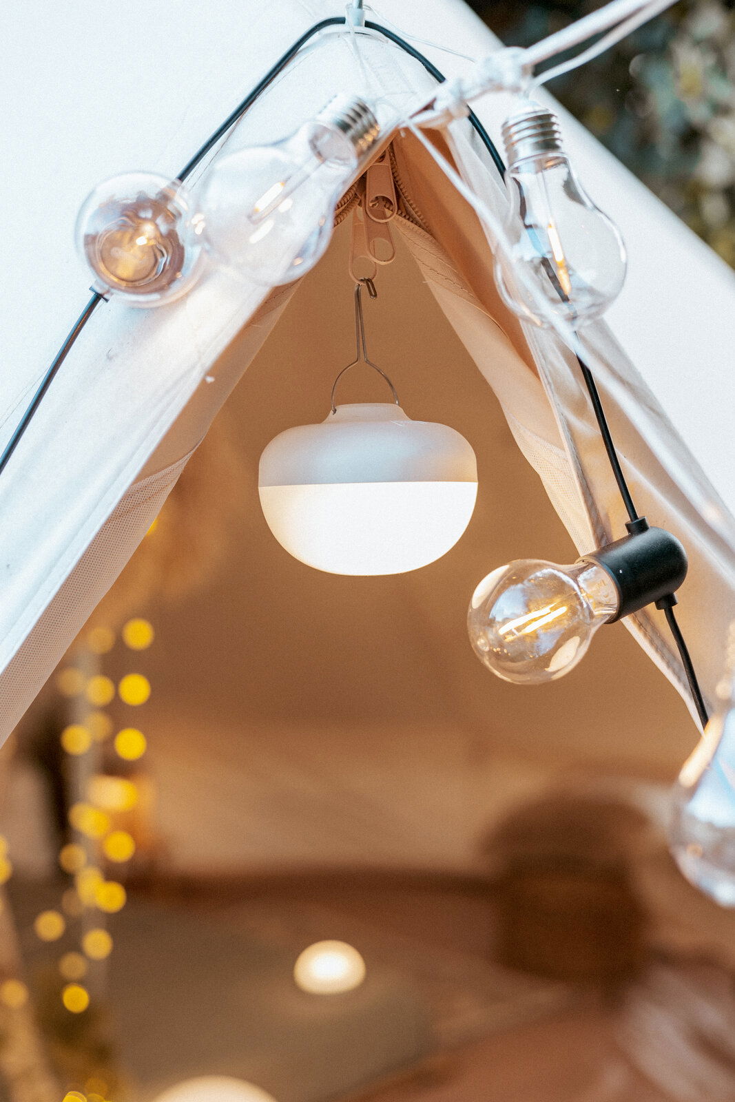 Nahaufnahme von oben auf den Zelteingang: eine runde, weiße Campinglampe hängt im Inneren, umgeben von einer Lichterkette mit Glühbirnen.