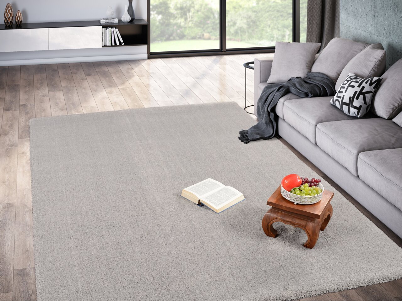 Grauer Teppich Soft Dream ca. 240x340cm in einem modernen Wohnzimmer mit grauem Sofa, Holzboden und einem kleinen Tisch mit Obstschale und Buch, aus der Vogelperspektive.