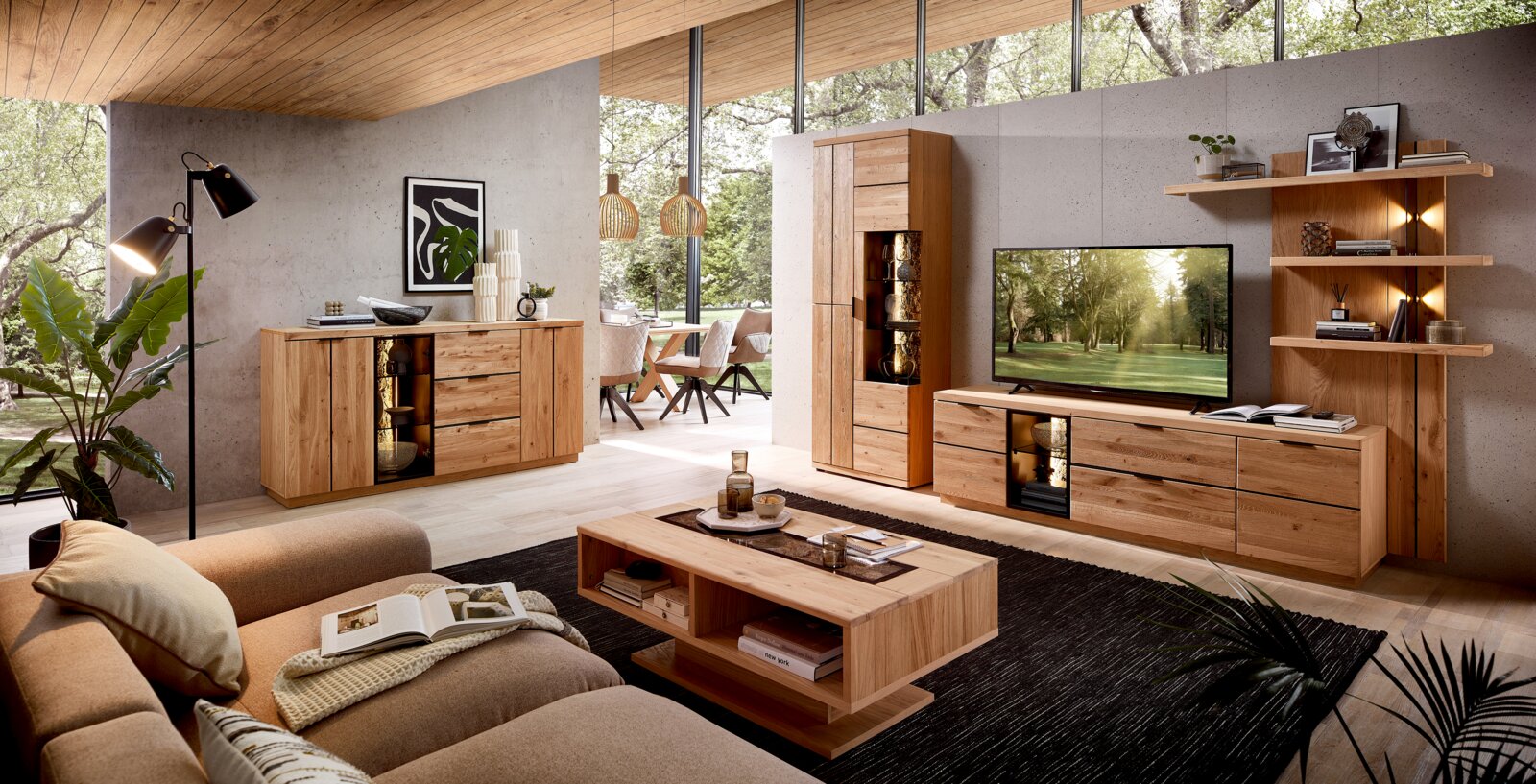 Modernes Wohnzimmer mit Wohnwand und Sideboard aus hellem Massivholz, TV-Lowboard und Vitrine mit Glas-/LED-Elementen sowie Couchtisch auf dunklem Teppich, perspektivische Raumansicht von schräg vorne