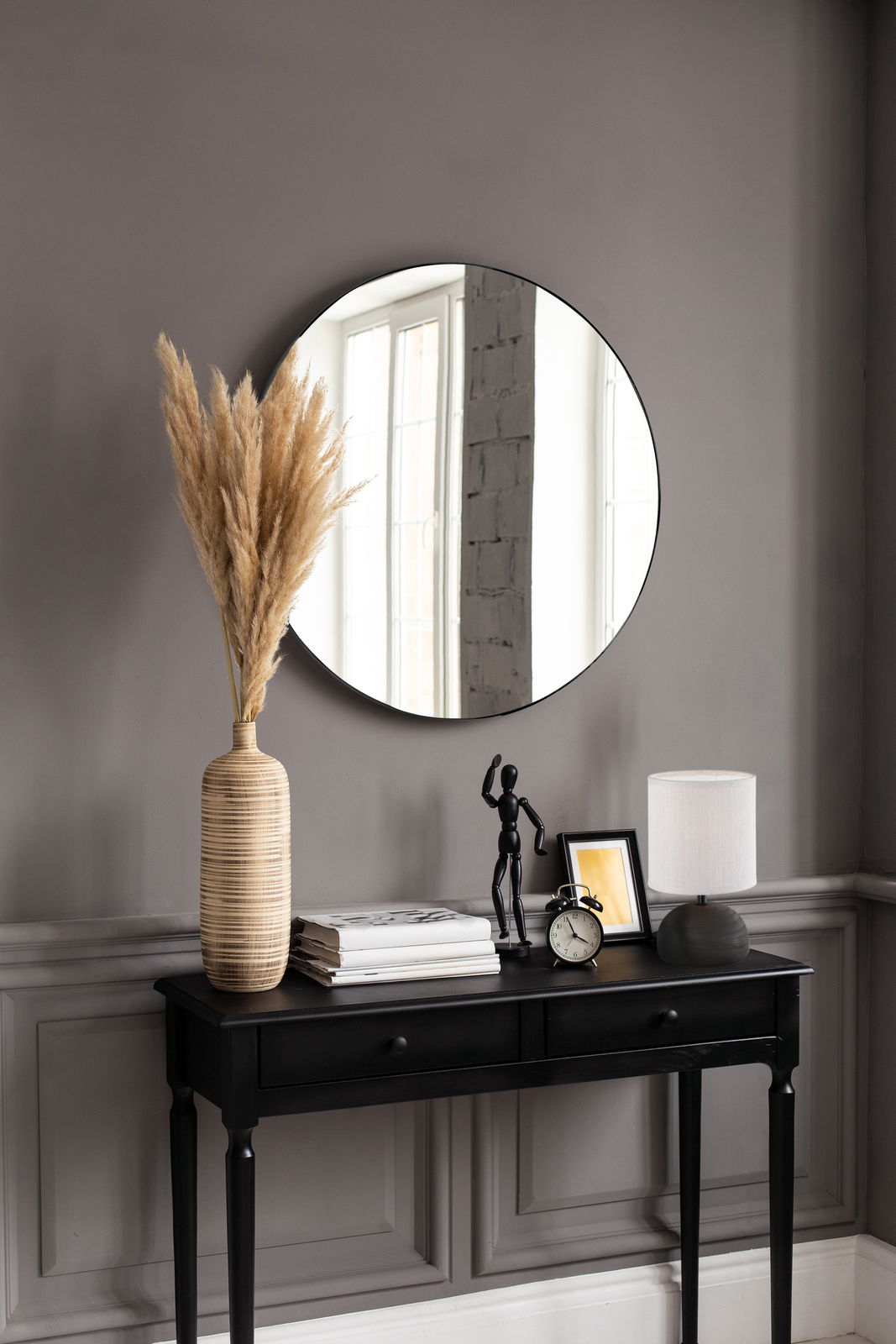 Runder Spiegel mit einem Durchmesser von ca. 50 cm, an einer grauen Wand über einem schwarzen Konsolentisch mit Dekorationen wie einer Vase mit Pampasgras, einer Tischlampe und einem Wecker. Perspektive: Frontalansicht.