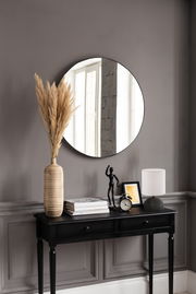 Runder Spiegel mit einem Durchmesser von ca. 50 cm, an einer grauen Wand über einem schwarzen Konsolentisch mit Dekorationen wie einer Vase mit Pampasgras, einer Tischlampe und einem Wecker. Perspektive: Frontalansicht.