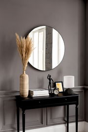 Runder Spiegel mit einem Durchmesser von ca. 50 cm, an einer grauen Wand über einem schwarzen Konsolentisch mit Dekorationen wie einer Vase mit Pampasgras, einer Tischlampe und einem Wecker. Perspektive: Frontalansicht.