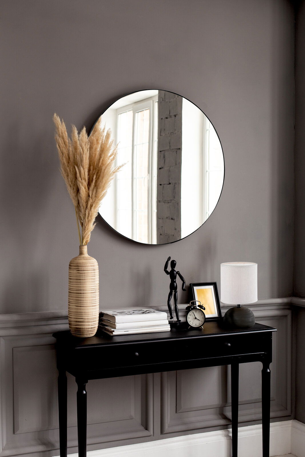 Runder Spiegel mit einem Durchmesser von ca. 50 cm, an einer grauen Wand über einem schwarzen Konsolentisch mit Dekorationen wie einer Vase mit Pampasgras, einer Tischlampe und einem Wecker. Perspektive: Frontalansicht.