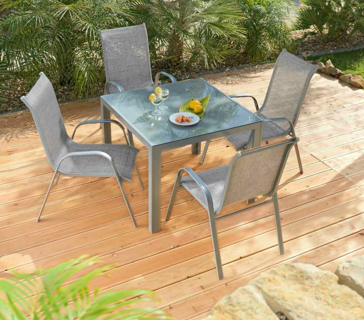 Moderner Gartentisch mit Glasplatte und vier grauen Stühlen auf einer Holzterrasse, umgeben von Pflanzen, aus der Vogelperspektive.