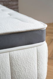 Nahaufnahme einer Matratze mit weißem, strukturiertem Stoff und grauem, atmungsaktivem Seitenstreifen, seitliche Perspektive