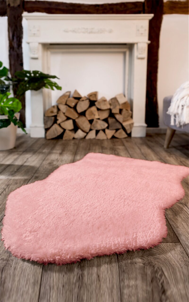Rosa Lammfell Teppich vor einem Kamin mit Holzstapeln, aus der Perspektive des Bodens aufgenommen.
