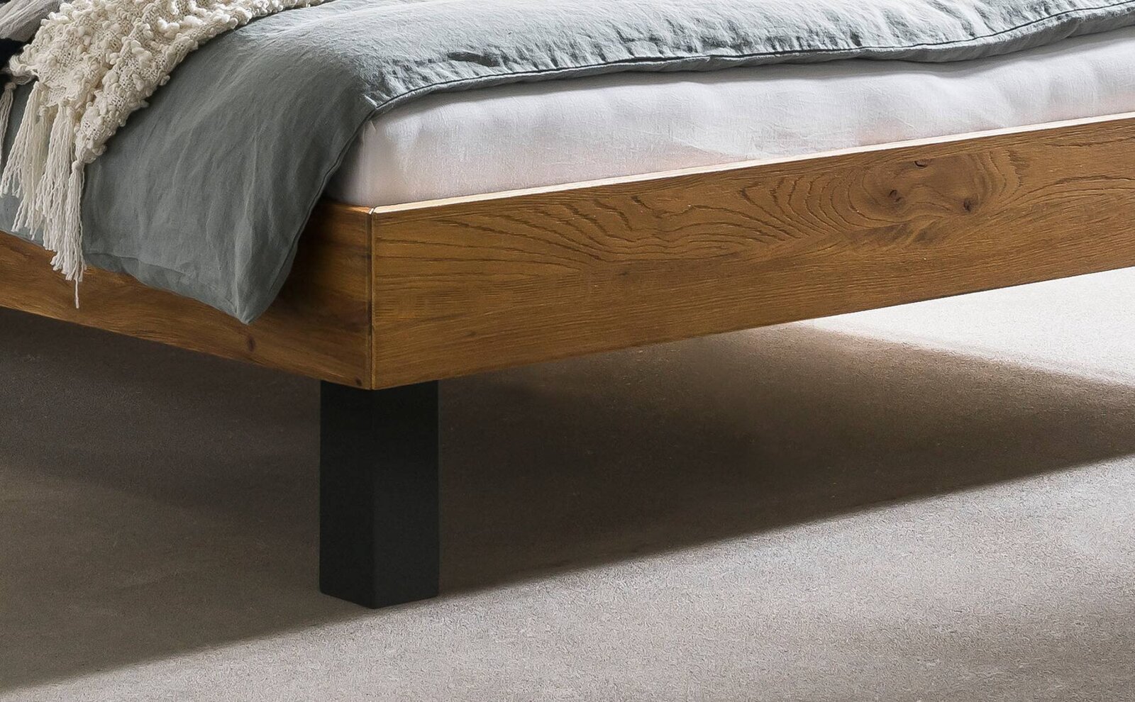Detailansicht eines Bettrahmens aus massivem Holz mit sichtbarer Maserung und schwarzem Metallfuß, mit grauer Bettwäsche und weißer Matratze, fotografiert in schräger Seitenansicht von vorne unten