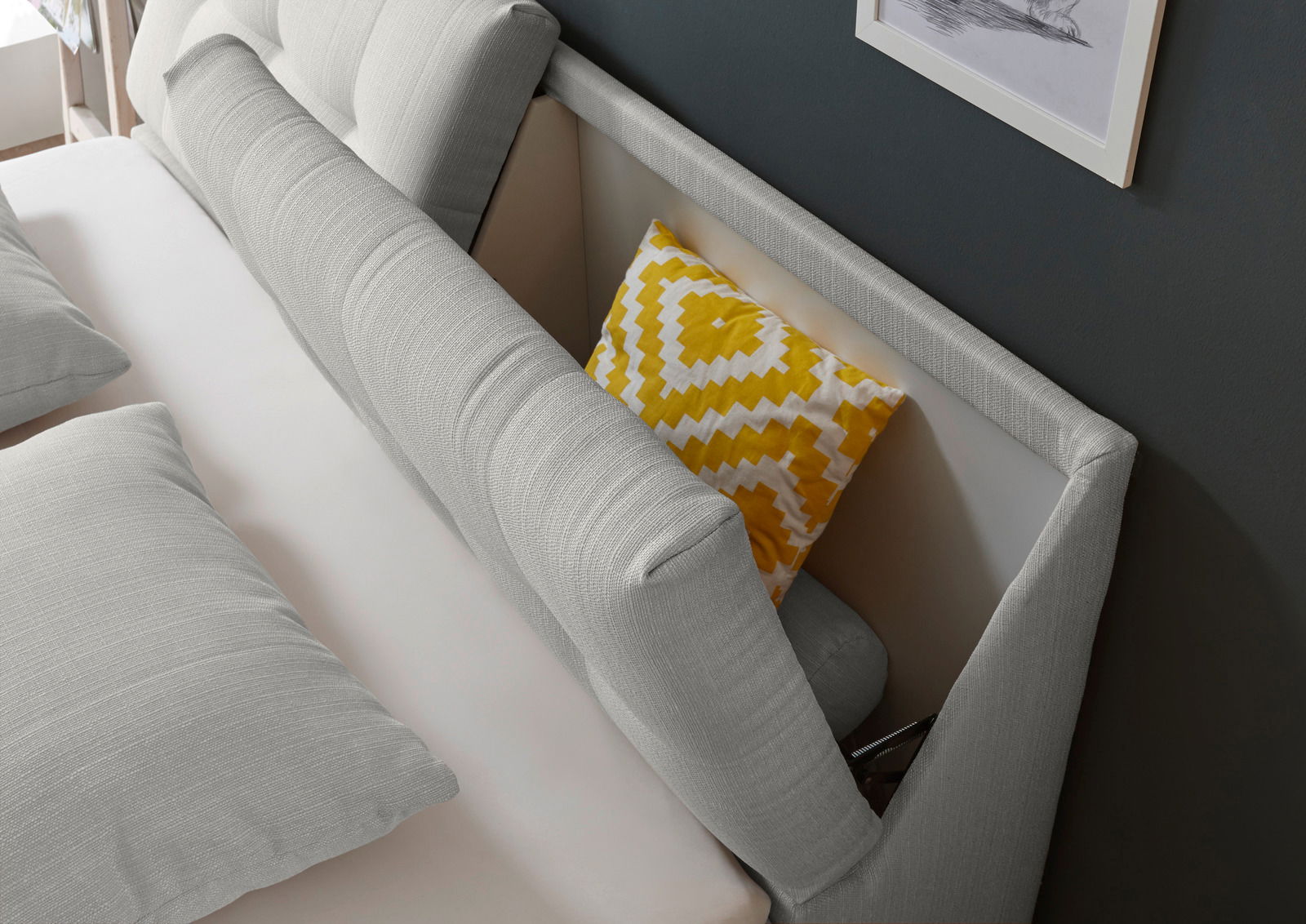 Nahaufnahme eines Boxspringbettes mit grauem Kopfteil und gelbem Zierkissen, seitliche Perspektive.