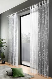 Transparenter Schlaufenschal mit floralem Muster, aufgehängt vor einem großen Fenster mit Blick auf die Stadt. Perspektive von innen, zeigt auch einen Teil des Wohnzimmers mit Pflanzen und Kissen.