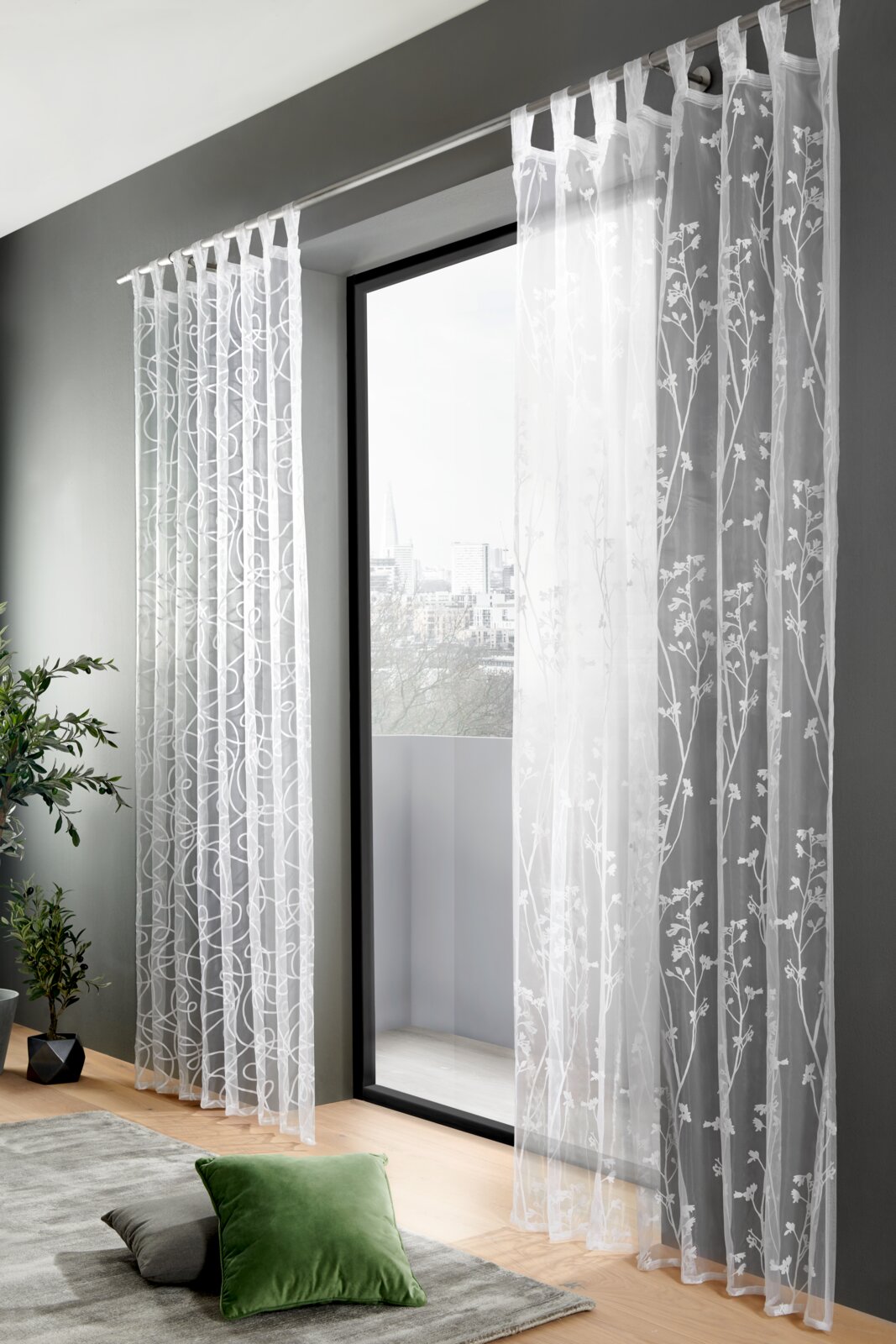 Transparenter Schlaufenschal mit floralem Muster, aufgehängt vor einem großen Fenster mit Blick auf die Stadt. Perspektive von innen, zeigt auch einen Teil des Wohnzimmers mit Pflanzen und Kissen.