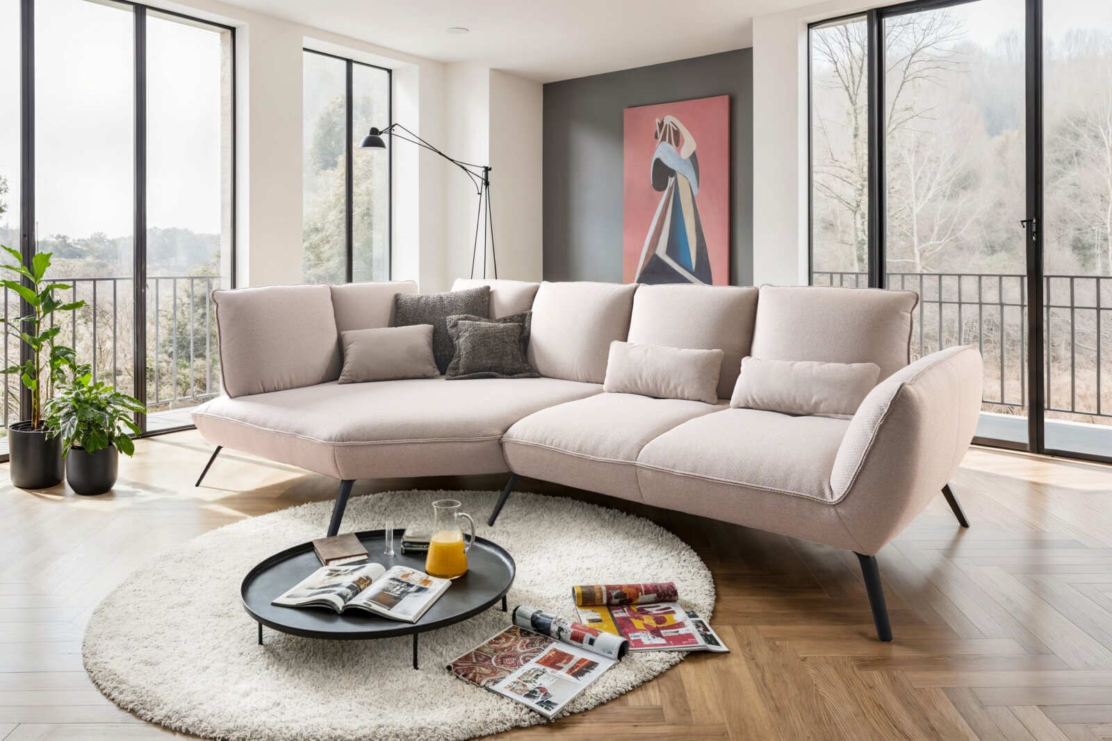 Moderne Longchairkombination in einem hellen Wohnzimmer mit großen Fenstern, die eine Aussicht auf die Natur bieten. Die Perspektive zeigt die Couch von vorne, umgeben von einem runden Teppich und einem Couchtisch mit Zeitschriften und einem Krug Orangens