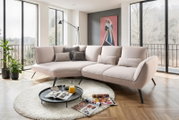 Moderne Longchairkombination in einem hellen Wohnzimmer mit großen Fenstern, die eine Aussicht auf die Natur bieten. Die Perspektive zeigt die Couch von vorne, umgeben von einem runden Teppich und einem Couchtisch mit Zeitschriften und einem Krug Orangens