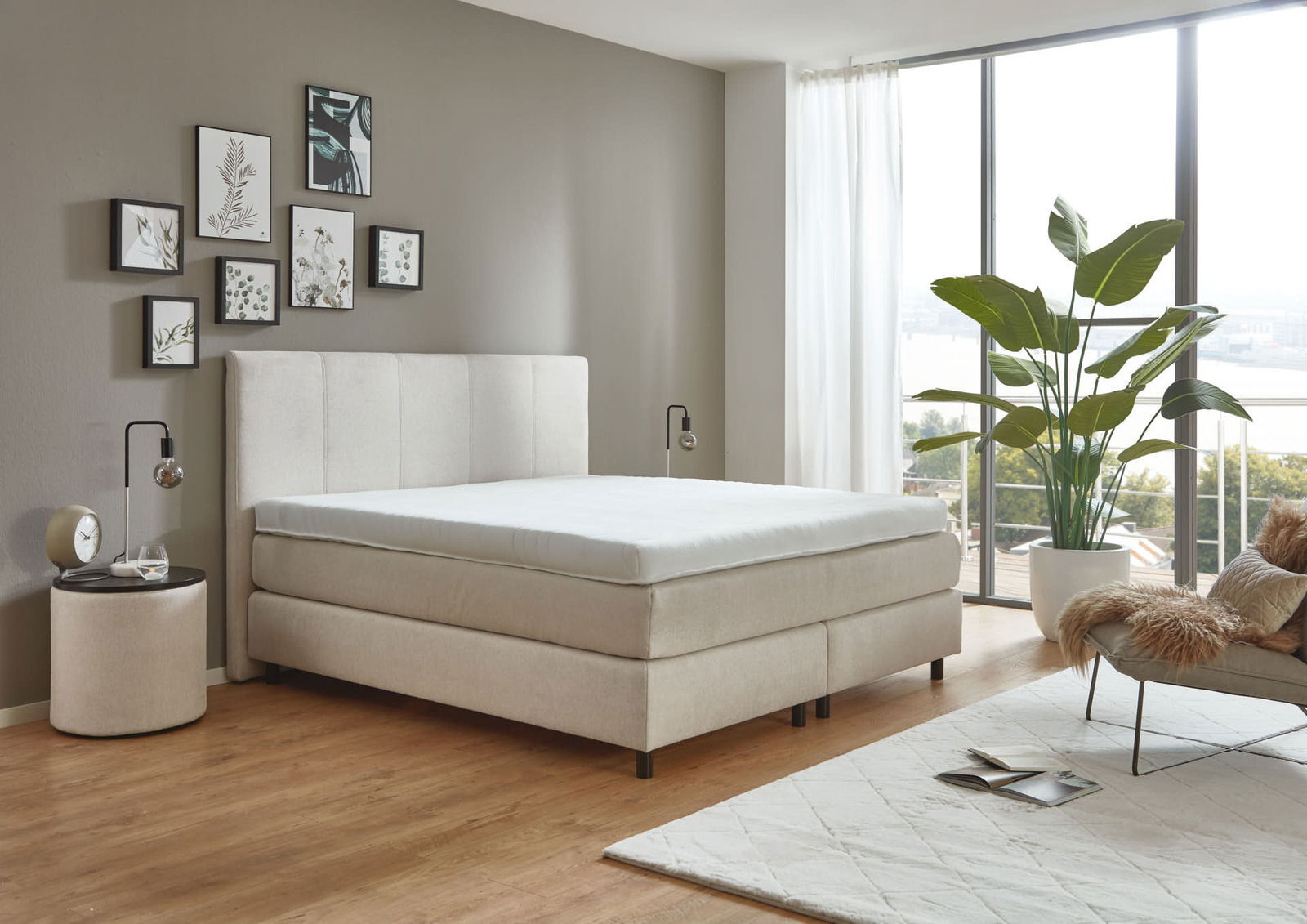 Modernes Boxspringbett in einem stilvollen Schlafzimmer mit großen Fenstern und Pflanzen, seitliche Perspektive.