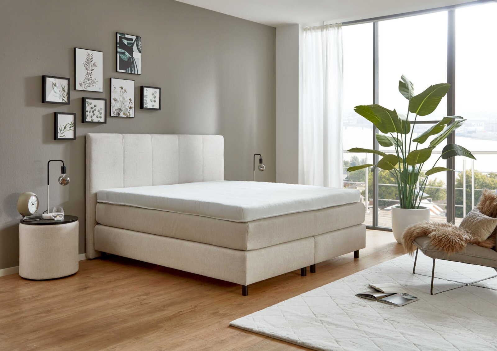 Modern boxspringbed in een stijlvolle slaapkamer met grote ramen en planten, zijaanzicht.