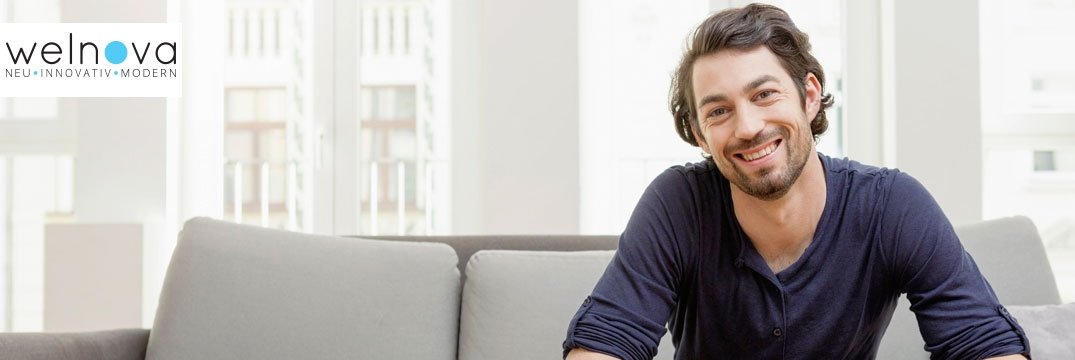 Ein lachender Mann sitzt auf einem grauen Sofa in einem hellen Wohnzimmer mit großen Fenstern.