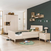 Kinderzimmer mit einem cremefarbenen Bett mit Absturzsicherung, umgeben von einem Schrank, einem Nachttisch und Dekorationen. Perspektive: seitliche Ansicht des Bettes.