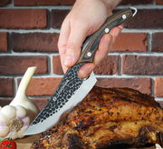 Kochmesser RANGER 15 cm mit gehämmertem Klingenmuster, Holzgriff und einer Hand, die es hält, während es ein gebratenes Stück Fleisch schneidet. Perspektive von der Seite.