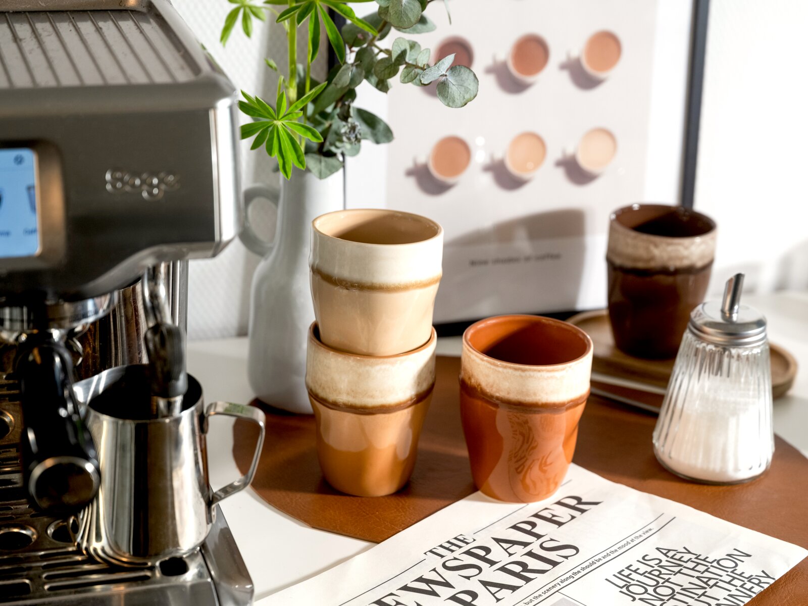 Stilvolles Keramikbecher-Set in warmen Braun- und Cremeglasuren auf einem Tablett neben Espressomaschine, Milchkanne und Zuckerdose, aufgenommen aus leicht schräger Frontperspektive