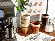Stilvolles Keramikbecher-Set in warmen Braun- und Cremeglasuren auf einem Tablett neben Espressomaschine, Milchkanne und Zuckerdose, aufgenommen aus leicht schräger Frontperspektive