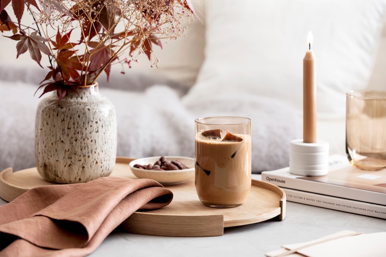 Like by Villeroy & Boch Set van 2 longdrinkbekers LIKE CLAY Longdrinkbecher mit Eiskaffee auf einem Holztablett, umgeben von einer Vase mit Herbstblättern, einer brennenden Kerze und einem Buch, aus der Seitenperspektive fotografiert.