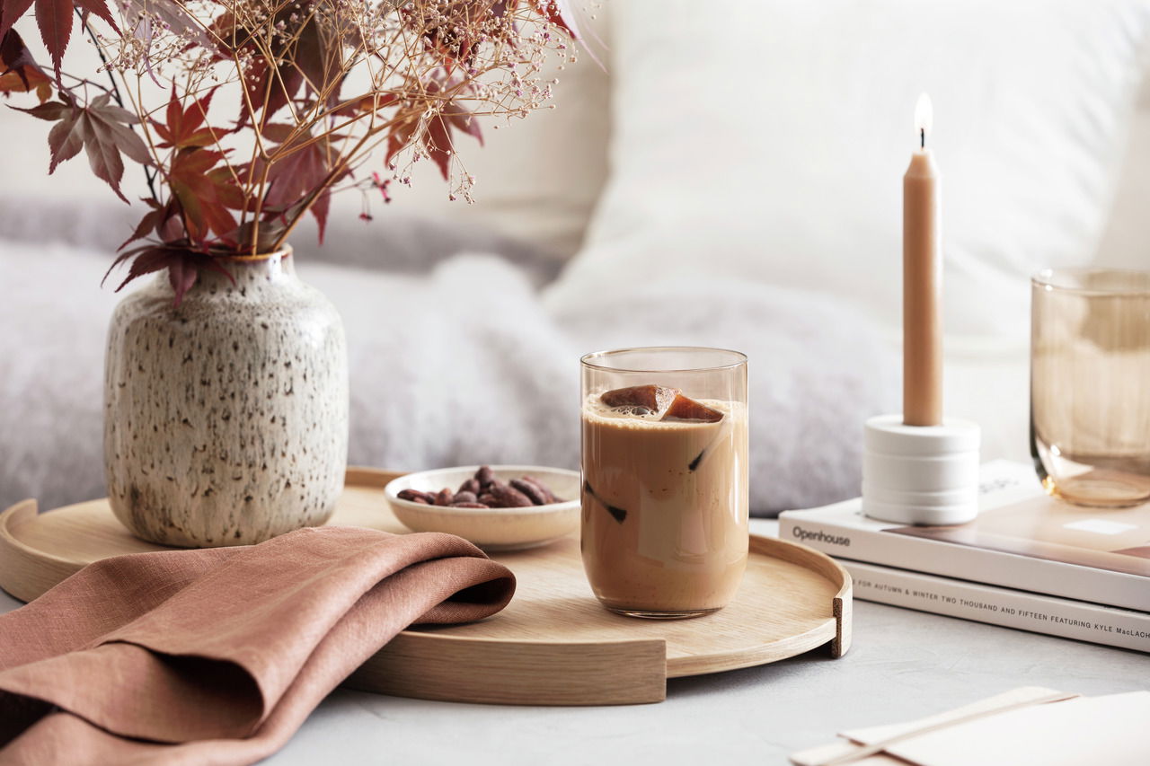 Longdrinkbecher mit Eiskaffee auf einem Holztablett, umgeben von einer Vase mit Herbstblättern, einer brennenden Kerze und einem Buch, aus der Seitenperspektive fotografiert.