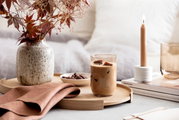 Longdrinkbecher mit Eiskaffee auf einem Holztablett, umgeben von einer Vase mit Herbstblättern, einer brennenden Kerze und einem Buch, aus der Seitenperspektive fotografiert.