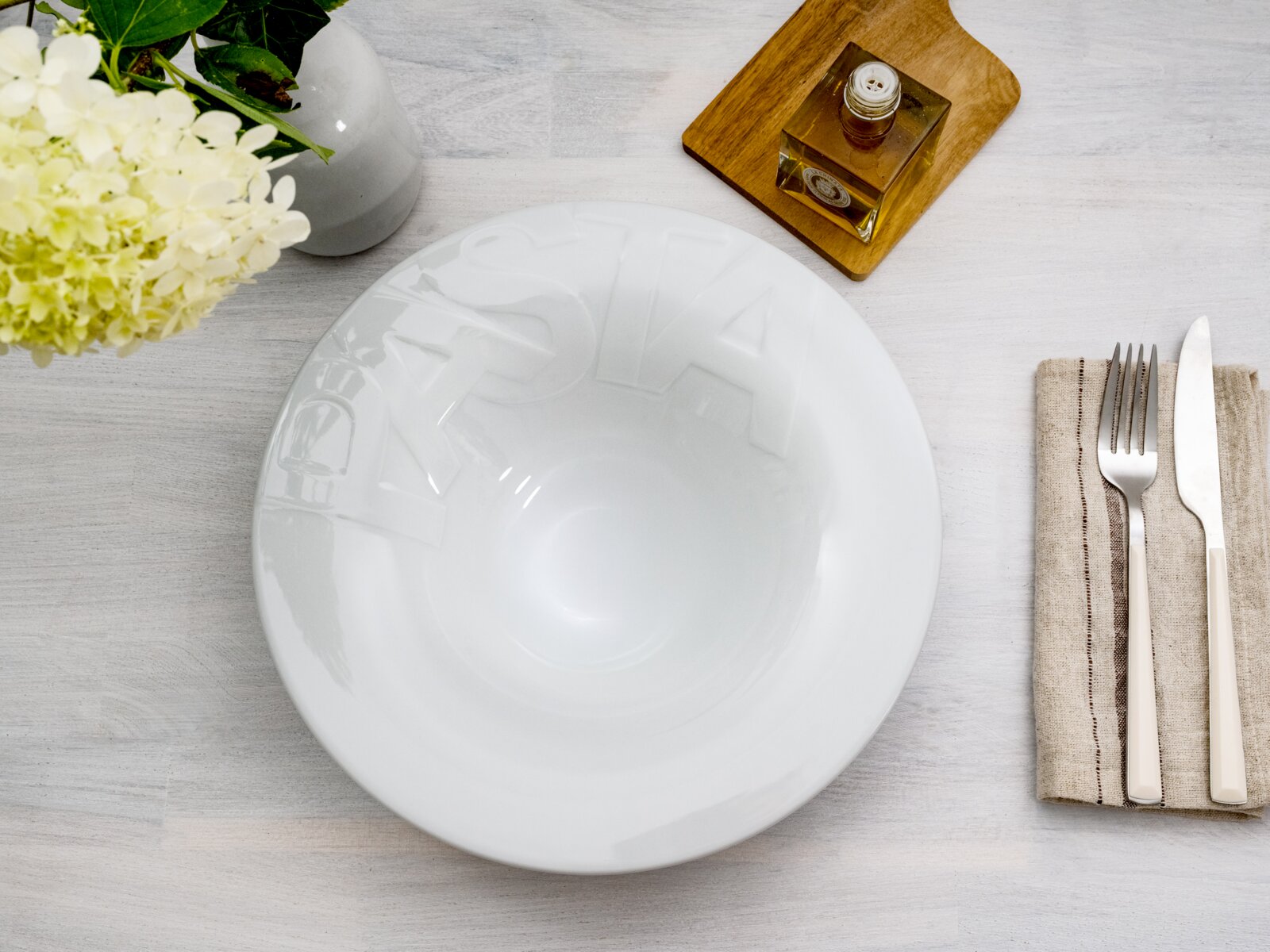 Weiße, glänzende Pastaschale mit erhabener Schrift „PASTA“, Draufsicht auf hellgrauem Holztisch; daneben Besteck auf Leinenserviette, Vase mit Blumen und Ölflasche auf Holzbrett.
