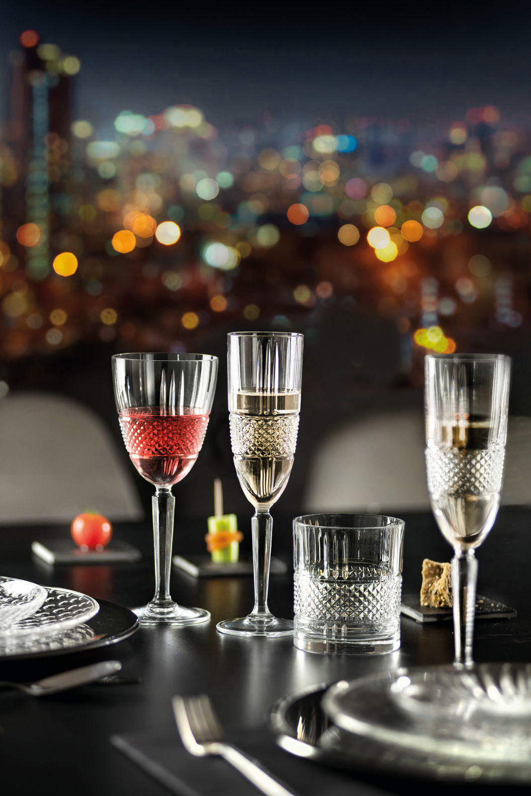 Ein elegantes Becherset mit verschiedenen Gläsern auf einem gedeckten Tisch, im Hintergrund eine verschwommene Stadtansicht bei Nacht. Perspektive: seitlich.