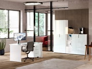 Modernes Büro mit weißem Schreibtisch und Rollcontainer im Vordergrund, Aktenschränken im Hintergrund, seitliche Perspektive.