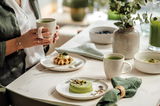 Frühstücksszene mit einem gedeckten Tisch, auf dem ein Frühstücksset mit Tassen, Tellern und Schalen zu sehen ist. Eine Person hält eine Tasse in der Hand. Perspektive von schräg oben.