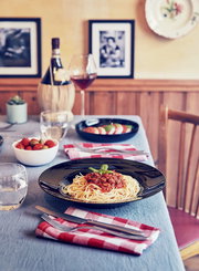 Schwarzer Pastateller mit Spaghetti Bolognese und Basilikum auf gedecktem Tisch mit Wein und Salat, Perspektive: leicht erhöhte Frontansicht