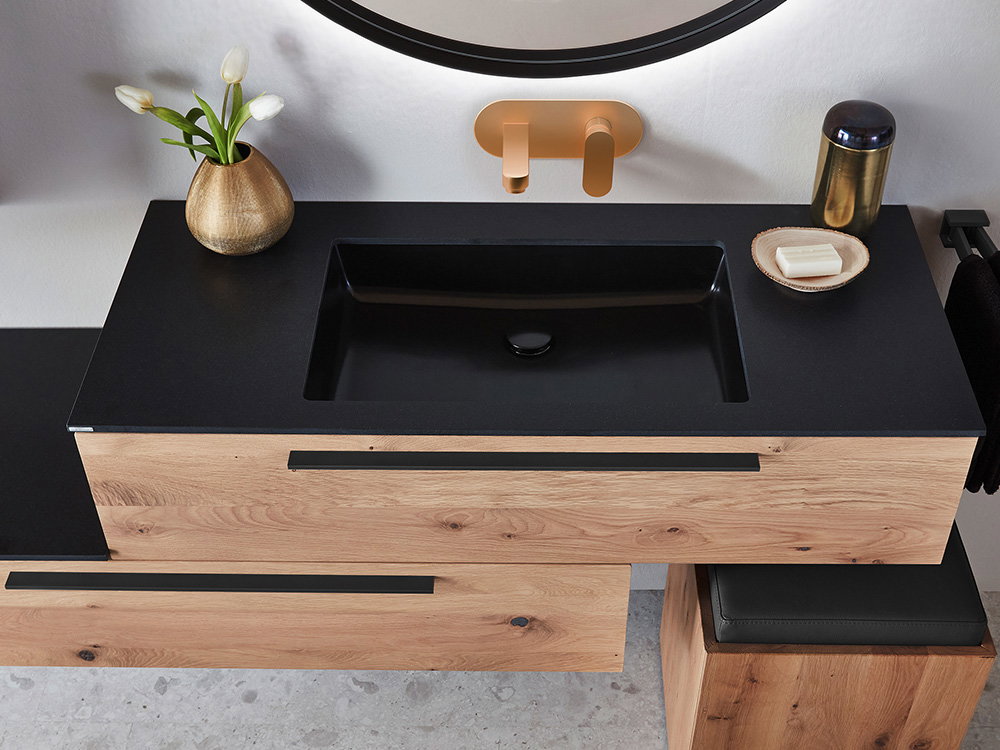 Holz-Waschtisch mit schwarzem Becken, goldener Armatur und Deko mit Blume und Seife.
